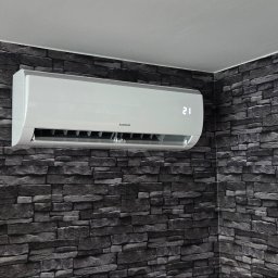 INSTALSERW - ROMAN RACZEK - Nowoczesna klimatyzacja ścienna KAISAI z cyfrowym wyświetlaczem '21', zamontowana w narożniku na tle ciemnej ściany imitującej kamień.