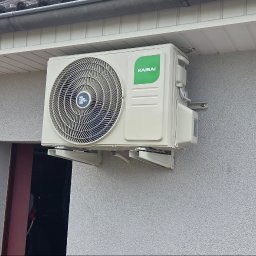 INSTALSERW - ROMAN RACZEK - Zewnętrzna jednostka klimatyzatora KAISAI na jasnoszarej, strukturalnej elewacji domu, pod ciemnym dachem z rynną i białą podbitką.