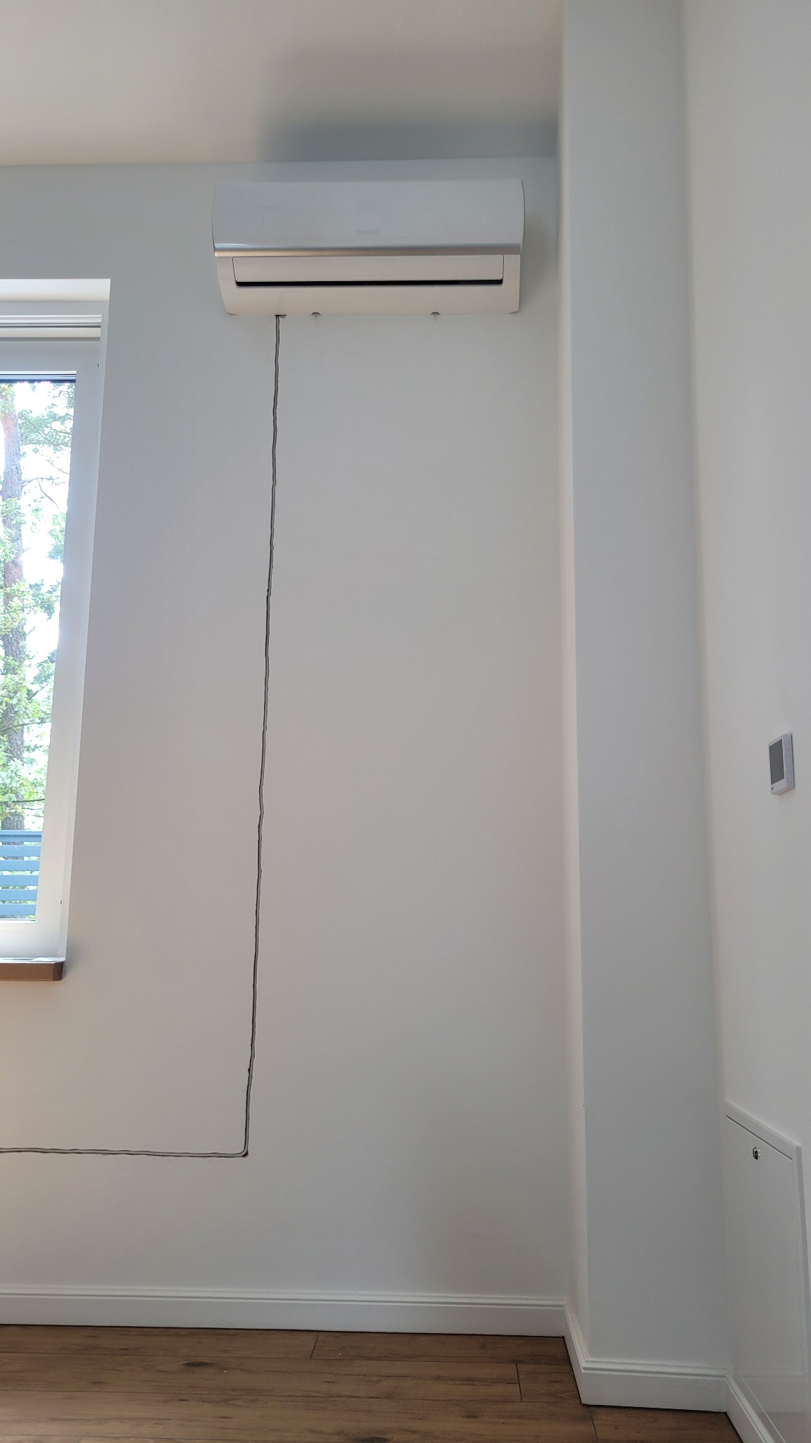 Biały klimatyzator ścienny zamontowany w jasnym pokoju z widocznym kablem zasilającym biegnącym wzdłuż ściany do listwy przypodłogowej.