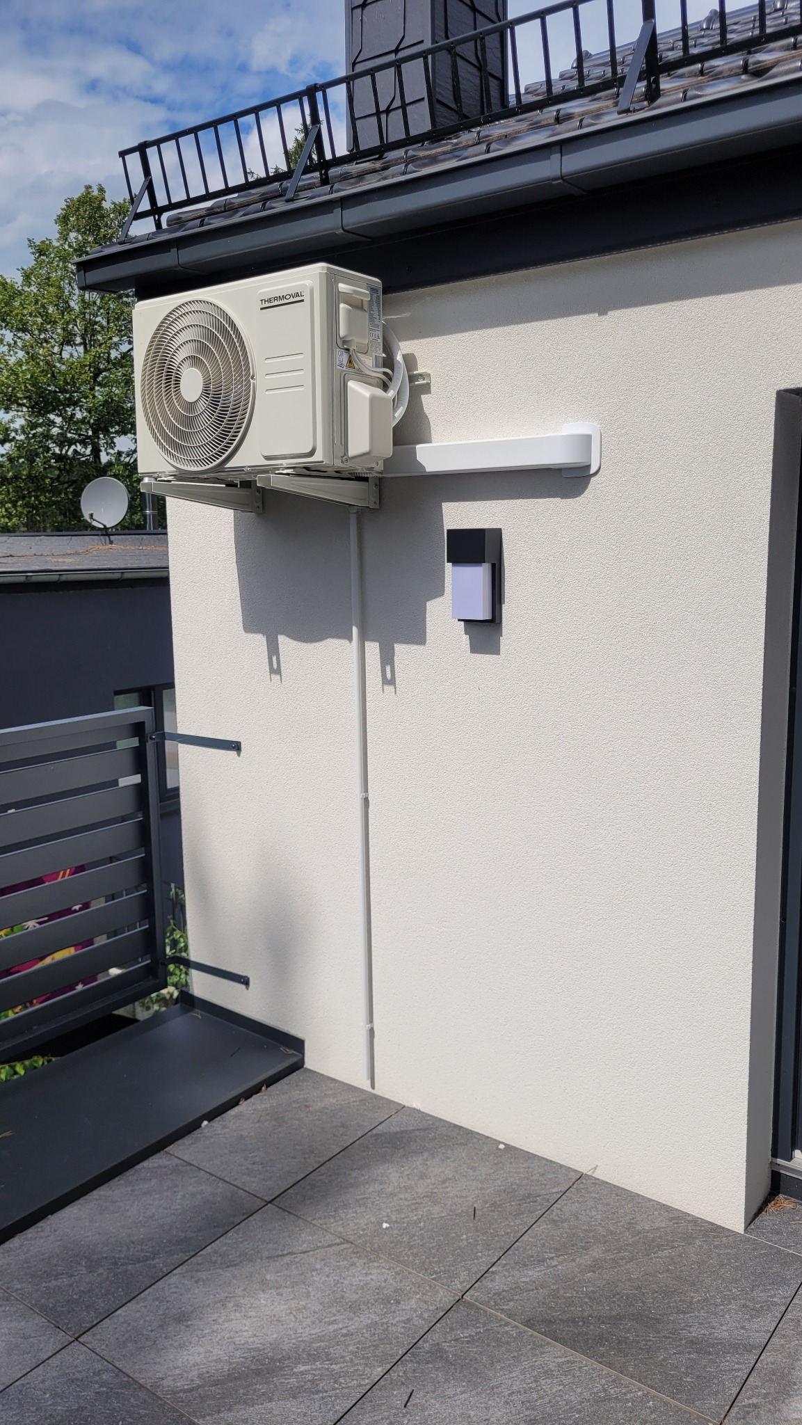 Klimatyzator Thermoval zamontowany na elewacji domu, widoczne przewody i listwa maskująca, balkon z szarymi płytkami w tle.
