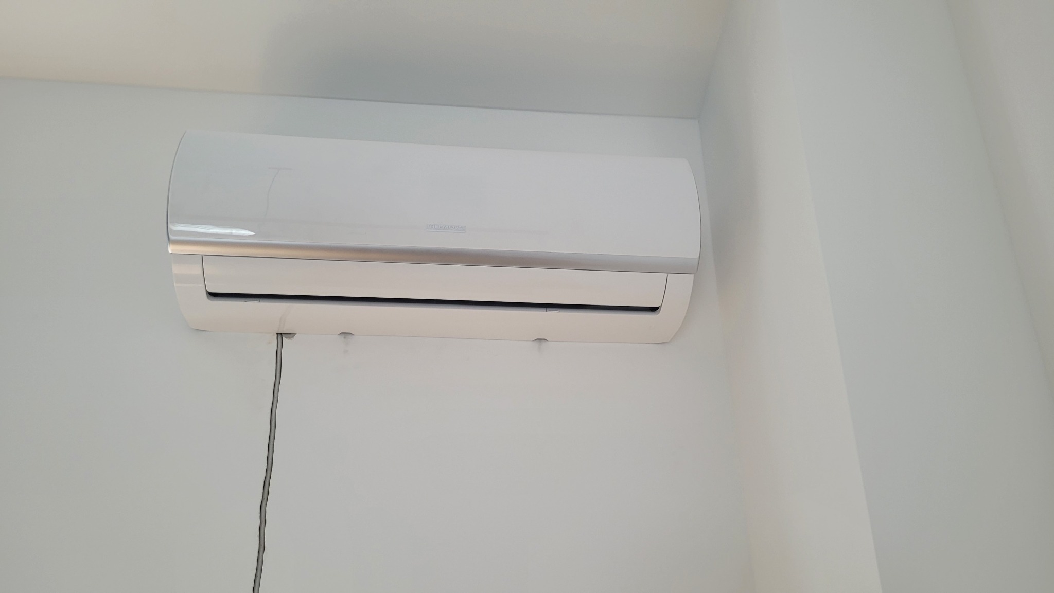 Biały klimatyzator ścienny marki Thermoval zamontowany w rogu białego pomieszczenia, widoczny kabel zasilający.