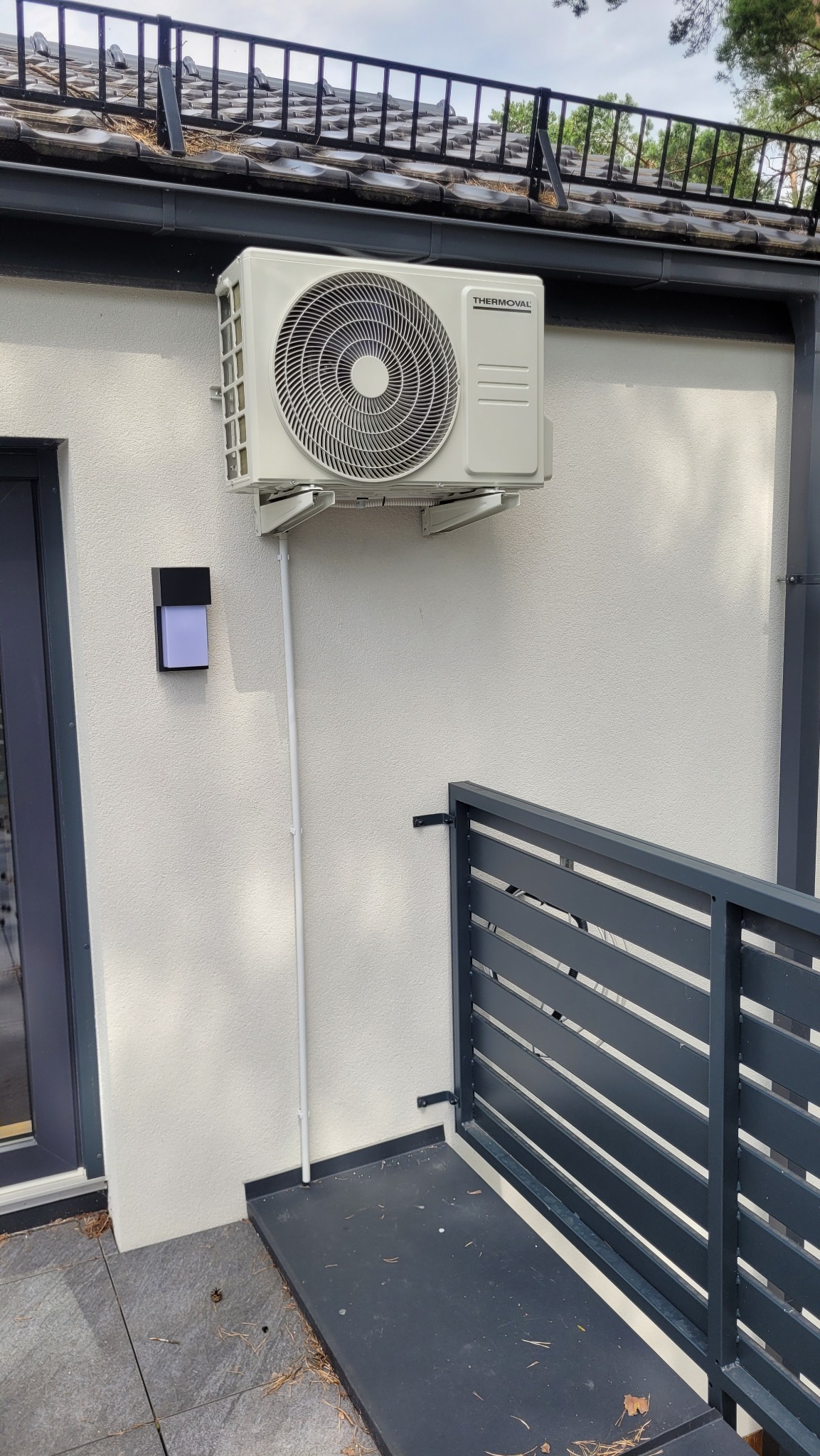 Zewnętrzna jednostka klimatyzacji marki Thermoval zamontowana na białej elewacji domu, widoczna rura odprowadzająca skropliny, obok czarna lampa ścienna i fragment szarej balustrady.