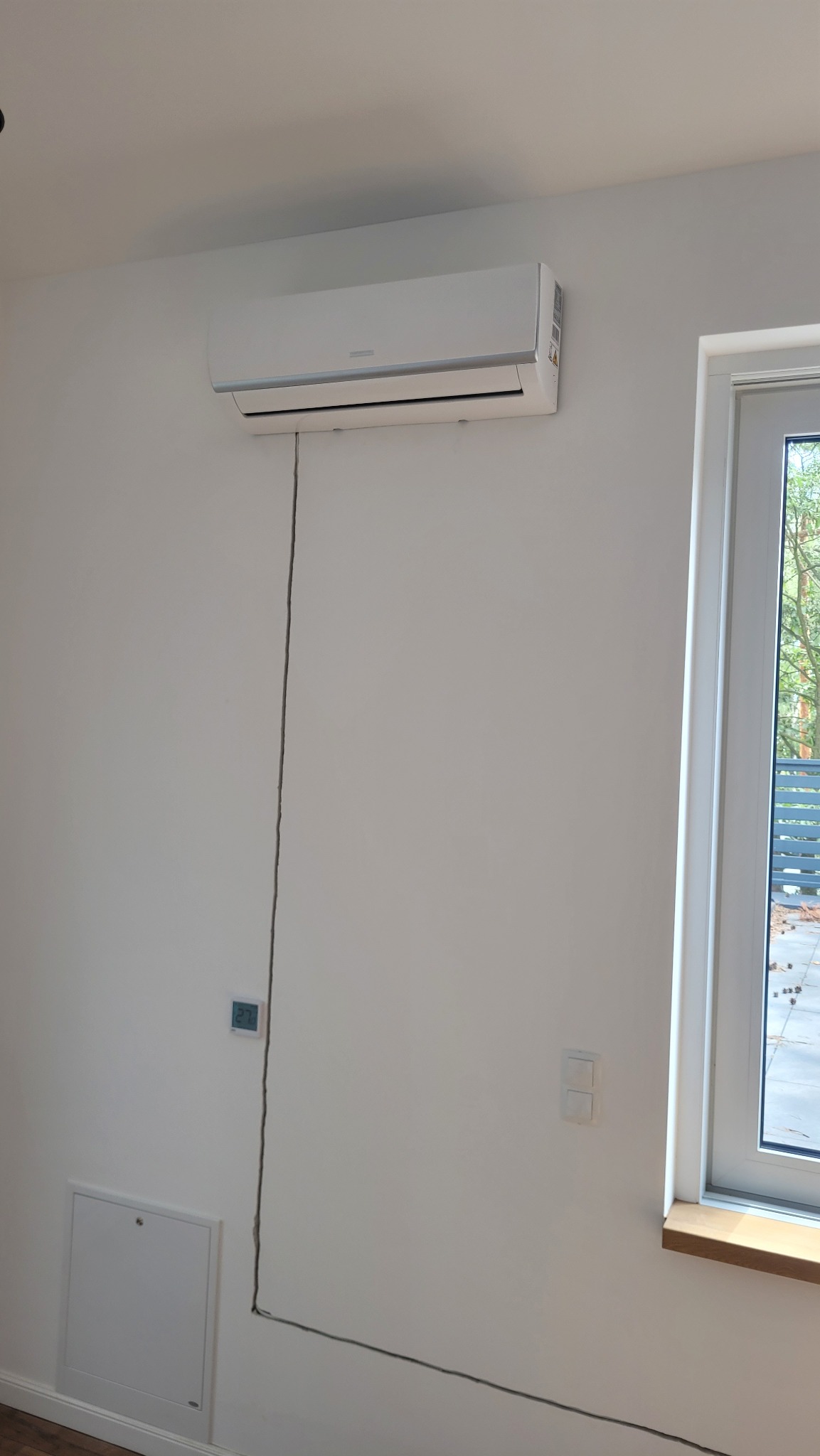 Biała klimatyzacja zamontowana na ścianie w jasnym pokoju, widoczny kabel zasilający biegnący wzdłuż ściany, okno z drewnianym parapetem po prawej stronie.