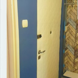 Maciej Rosiński - Białe, pikowane drzwi wejściowe z zamkiem i klamką, na tle granatowej ściany z włącznikiem światła i fragmentu ściany wyłożonej drewnopodobną płytką w jodełkę, widoczny mop i niebieski pojemnik.