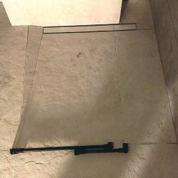 Maciej Rosiński - Ułożone, beżowe płytki podłogowe w łazience, widoczna czarna listwa odpływowa liniowa oraz narzędzie do cięcia płytek.