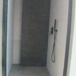 Maciej Rosiński - Wąska kabina prysznicowa z ciemnoszarą ścianą i czarną armaturą, widoczny odpływ liniowy w podłodze z szarych płytek.