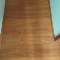 Maciej Rosiński - Nowo położona, jasnobrązowa podłoga z paneli drewnianych, widoczna z góry, z delikatnym wzorem słojów i niewielkimi niedoskonałościami, przy białej ścianie z kratką wentylacyjną.