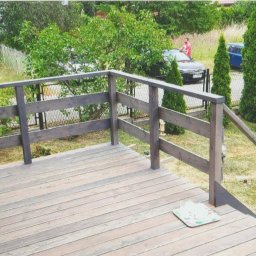 Maciej Rosiński - Drewniany taras z pomalowaną na szaro balustradą, widok na ogród z tujami, samochodem i osobą w tle.