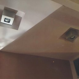 Maciej Rosiński - Sufit z dwoma kwadratowymi wnękami oświetleniowymi, jedna biała, druga w odcieniach brązu, z widocznymi lampami halogenowymi oraz fragment ściany pomalowanej na brązowo.