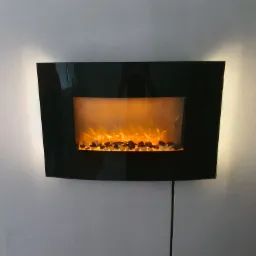 Czarny, elektryczny kominek ścienny z efektem płomieni i kamieniami dekoracyjnymi, oświetlony od tyłu, zamontowany na szarej ścianie. Widoczny kabel zasilający.