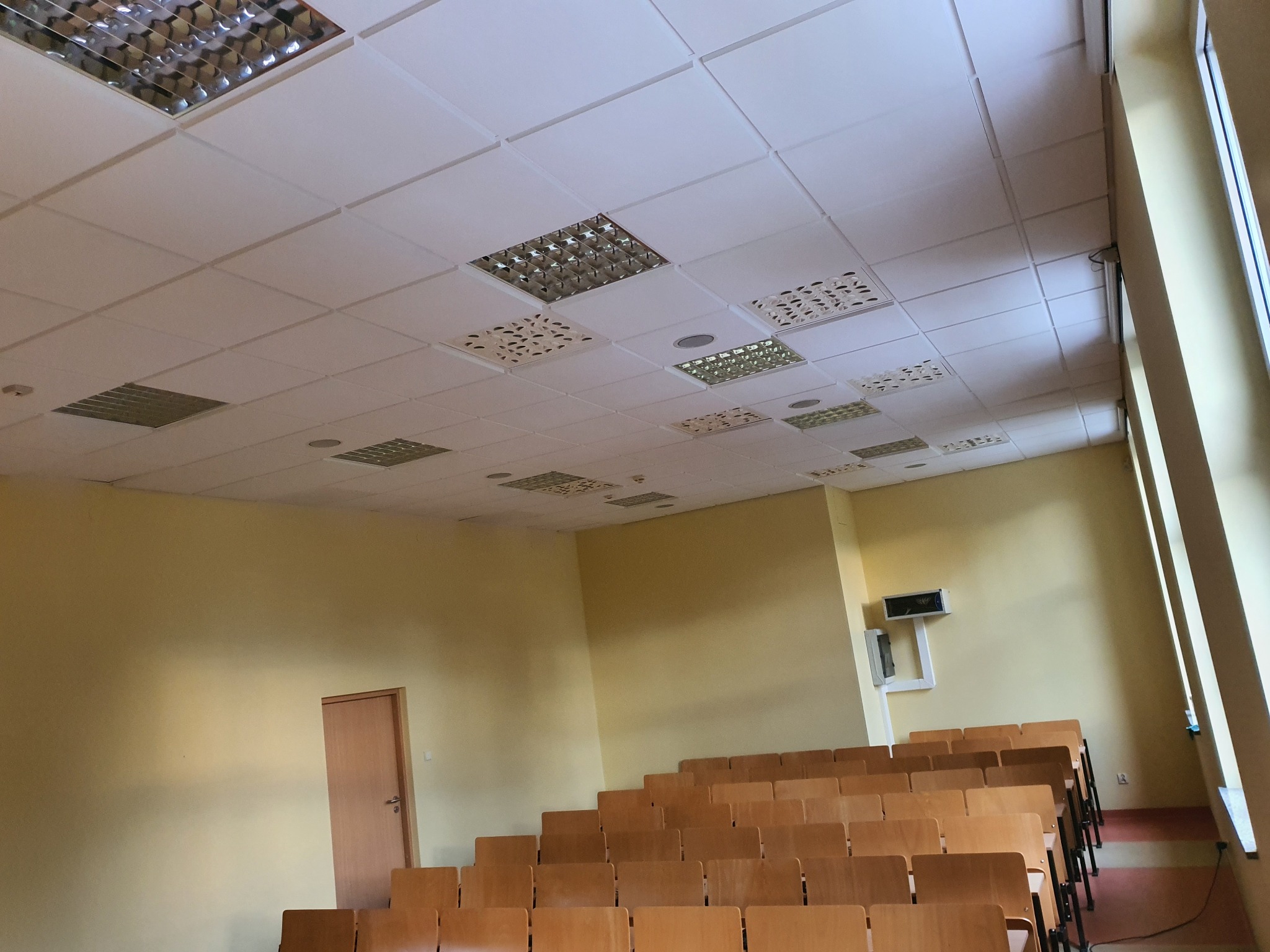 Sala wykładowa z rzędami drewnianych krzeseł, żółtymi ścianami i kasetonowym sufitem z różnymi wzorami opraw oświetleniowych.