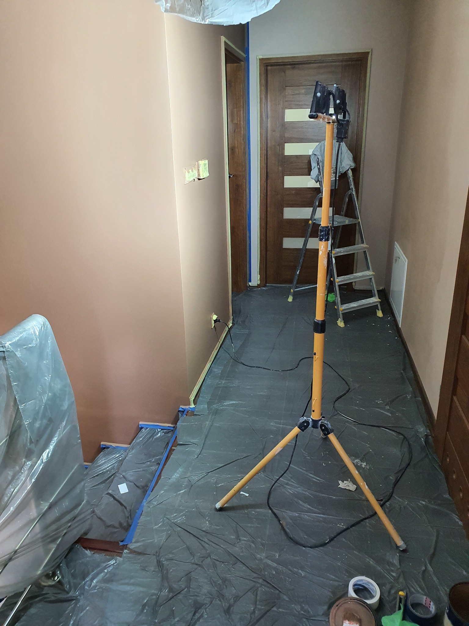 Korytarz w trakcie prac przygotowawczych: ściany częściowo pomalowane na brzoskwiniowy odcień, podłoga i schody zabezpieczone folią malarską, lampa na statywie oświetla przestrzeń, widoczne taśmy...