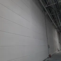 Wnętrze hali produkcyjnej z widoczną białą, panelową ścianą, instalacjami pod sufitem i regałami magazynowymi w tle.