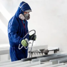 Pracownik w kombinezonie ochronnym i masce maluje metalowe profile natryskowo, używając pistoletu lakierniczego. Widoczne zielone rękawice ochronne i elementy instalacji natryskowej.