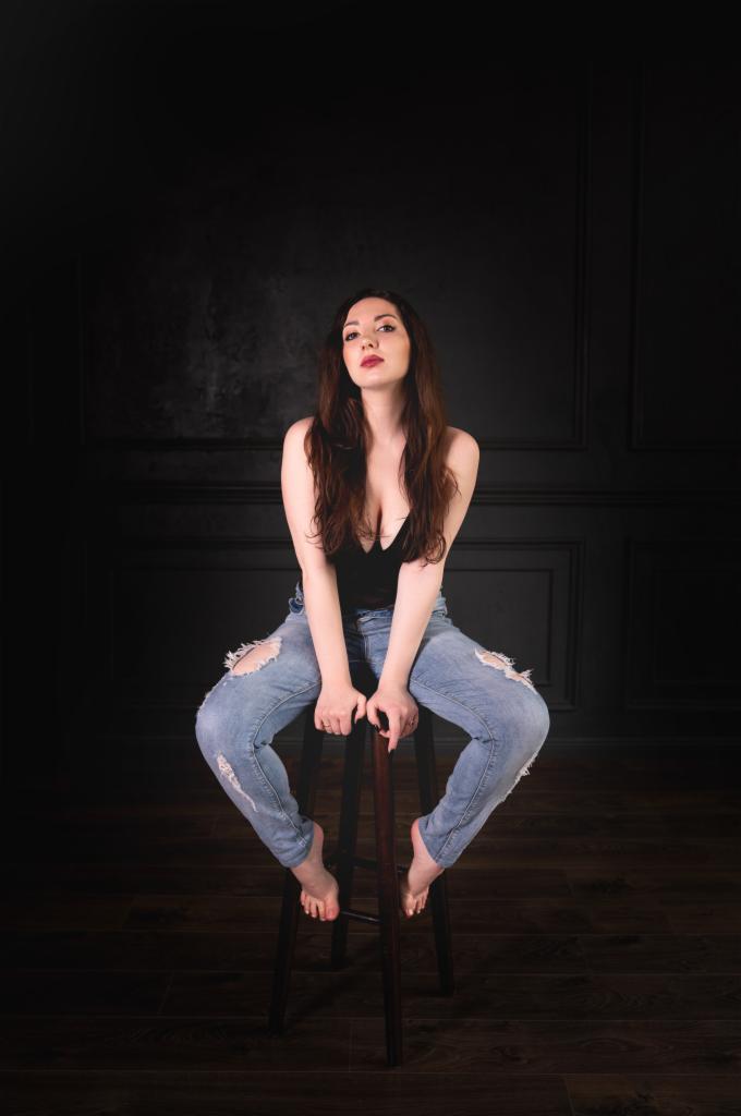Portret kobiety siedzącej na stołku w podartych dżinsach, czarnym topie i boso na tle ciemnej ściany z panelami.