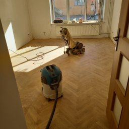 DANIEL DOMAŃSKI - Parkiet jodełka po cyklinowaniu, widoczna szlifierka do podłóg i odkurzacz przemysłowy Makita, pokój w trakcie prac wykończeniowych, naturalne światło z okna.