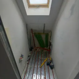 Montaż ogrzewania podłogowego w trakcie remontu poddasza z oknem dachowym; widoczne rury grzewcze, poziomica i wiertarka.