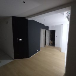 DANIEL DOMAŃSKI - Wnętrze mieszkania po świeżym malowaniu ścian na kolor antracytowy, z jasną drewnianą podłogą i widoczną instalacją elektryczną oraz drabina w przejściu do kolejnego pomieszczenia.