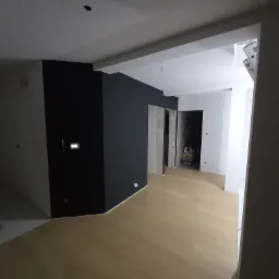 Wnętrze mieszkania po świeżym malowaniu ścian na kolor antracytowy, z jasną drewnianą podłogą i widoczną instalacją elektryczną oraz drabina w przejściu do kolejnego pomieszczenia.