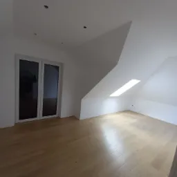 Puste, jasne poddasze z oknem dachowym, świeżo położoną parkietową podłogą i pomalowanymi na biało ścianami, widoczne otwory w suficie na oświetlenie.