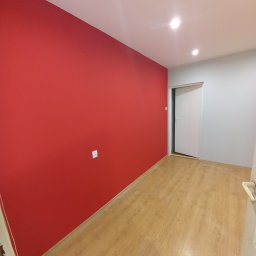 DANIEL DOMAŃSKI - Wnętrze pokoju po remoncie: jedna ściana pomalowana na intensywny czerwony kolor, pozostałe ściany szare, jasna drewniana podłoga, białe drzwi przesuwne w głębi, widoczny kontakt elektryczny.
