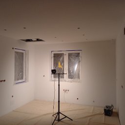 DANIEL DOMAŃSKI - Przygotowane do malowania pomieszczenie z zakrytymi folią oknami, zabezpieczoną podłogą, widocznymi puszkami elektrycznymi i lampą na statywie.