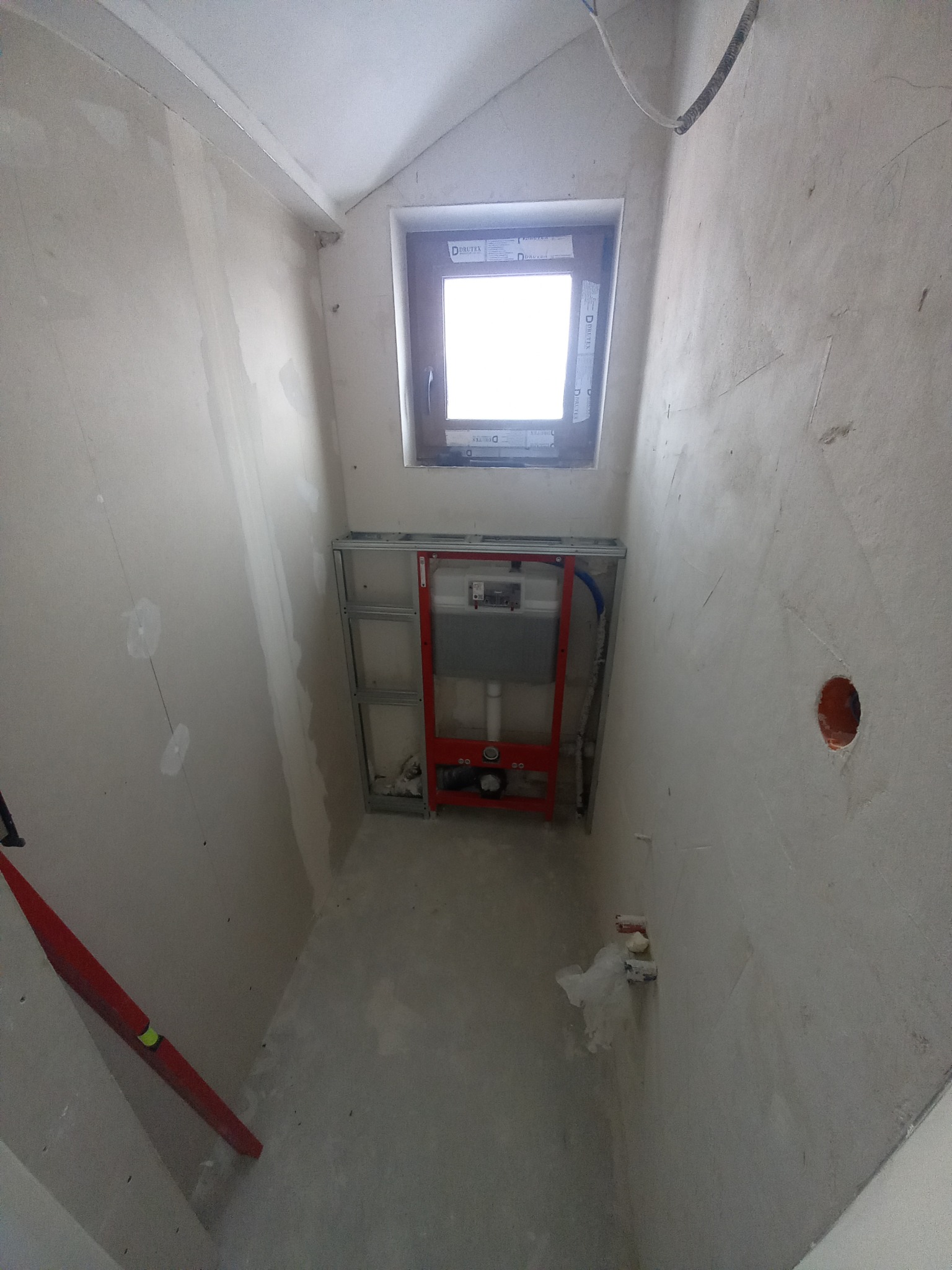 Wnętrze łazienki w trakcie prac przygotowawczych, widoczny stelaż podtynkowy WC na tle surowych, zagruntowanych ścian, okno i poziomica oparta o ścianę.