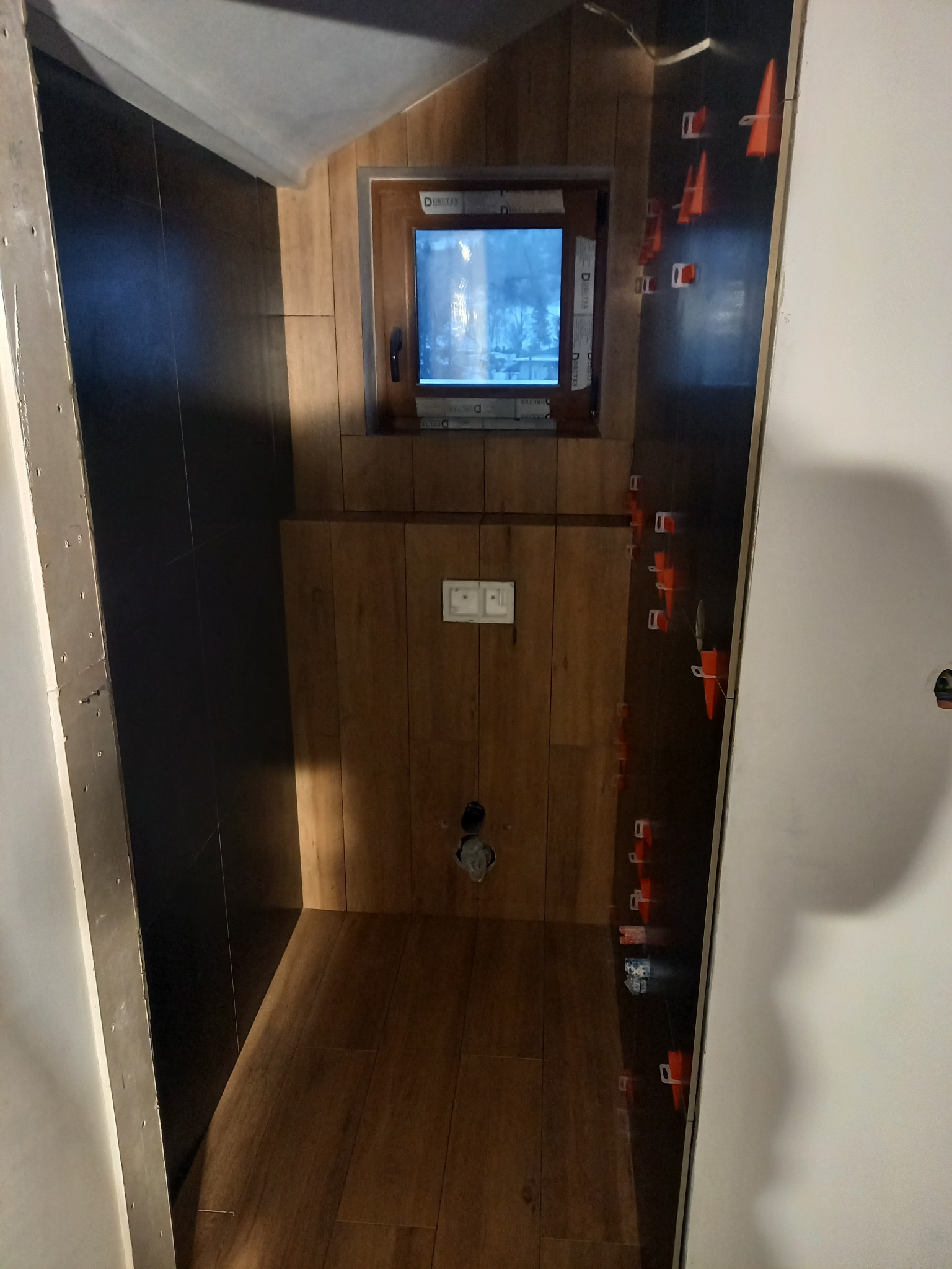 Wnętrze łazienki w trakcie układania płytek, widoczne czarne płytki na ścianach i drewnopodobne na podłodze oraz ścianie, z użyciem pomarańczowych klinów dystansowych, małe okno z widokiem...