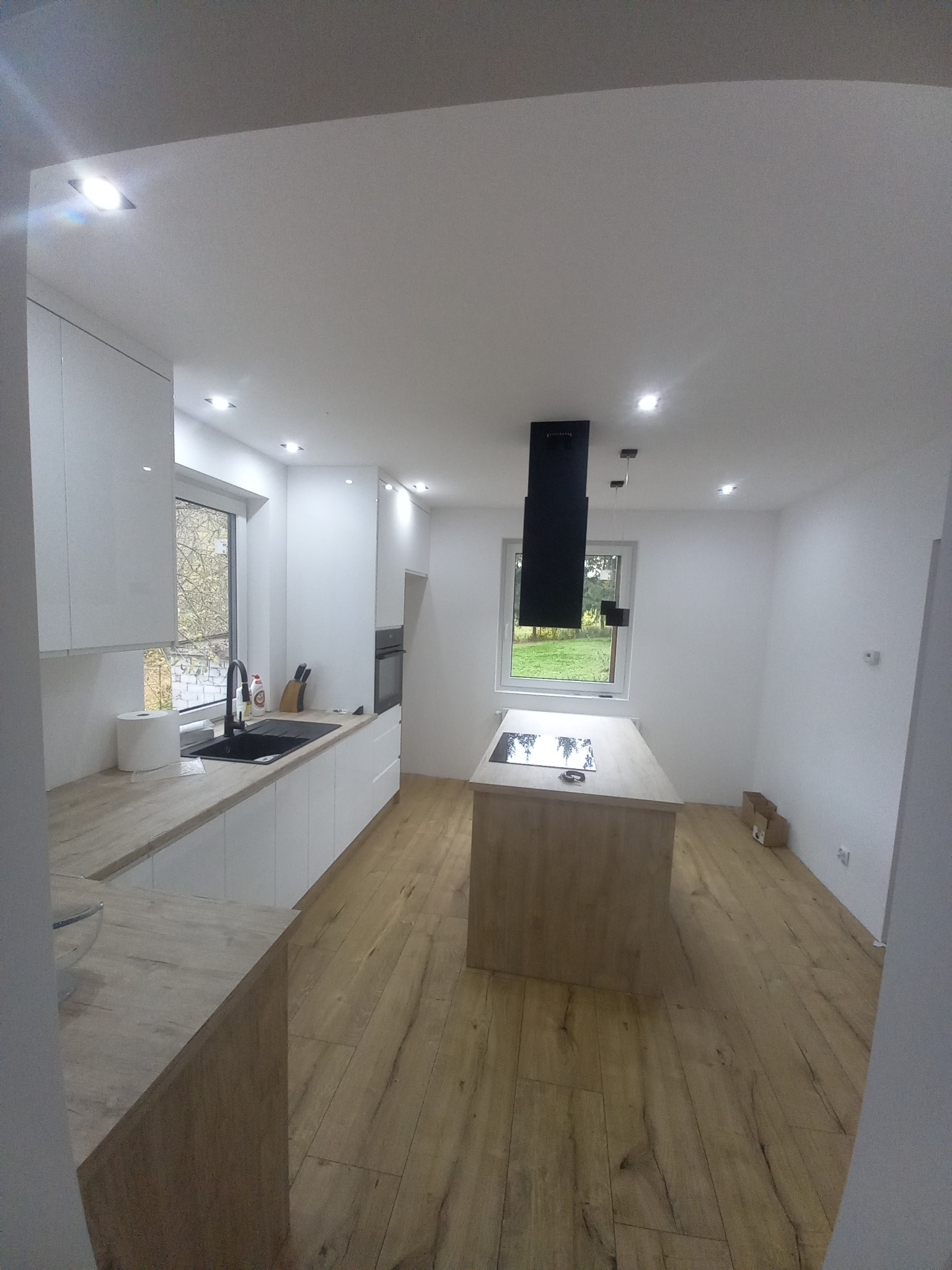 Nowoczesna kuchnia z białymi szafkami, drewnianym blatem i wyspą kuchenną z płytą indukcyjną, widok na ogród przez okno.