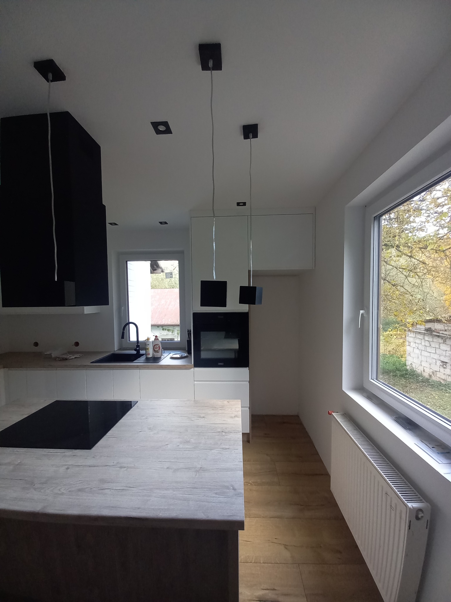 Nowoczesna kuchnia w bieli i czerni z drewnianym blatem wyspy, minimalistycznymi lampami i widokiem na ogród przez okno.