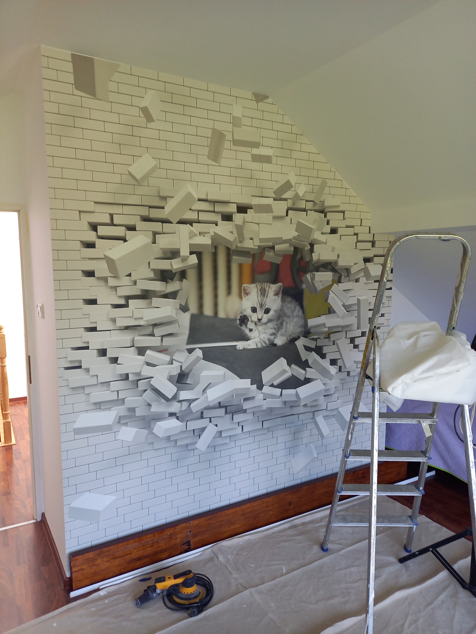 Ściana z fototapetą przedstawiającą białą cegłę z efektem 3D i wizerunkiem kociaka, drabina malarska, szlifierka oscylacyjna, zabezpieczona podłoga w trakcie prac.