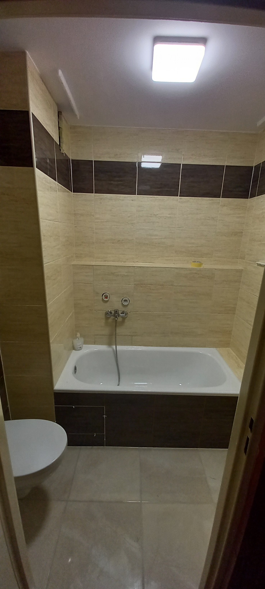 Wykończona łazienka z beżowymi i brązowymi płytkami, białą wanną oraz toaletą widoczną z boku, oświetlona kwadratową lampą sufitową.