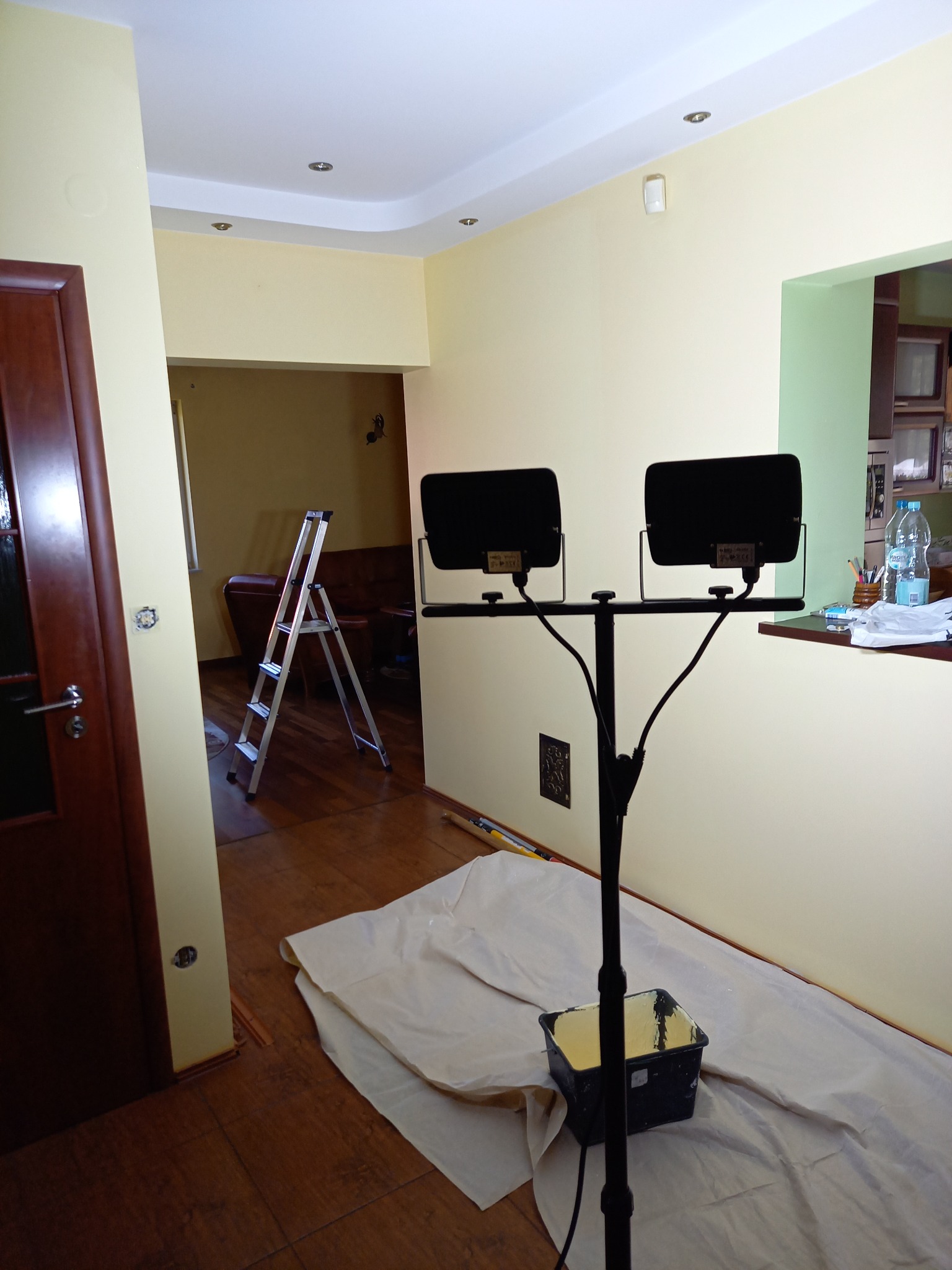 Przygotowania do prac wykończeniowych w jasnym pomieszczeniu: rozłożona folia ochronna na drewnianej podłodze, przenośny reflektor na statywie i drabina aluminiowa w przejściu do pokoju z brązową...