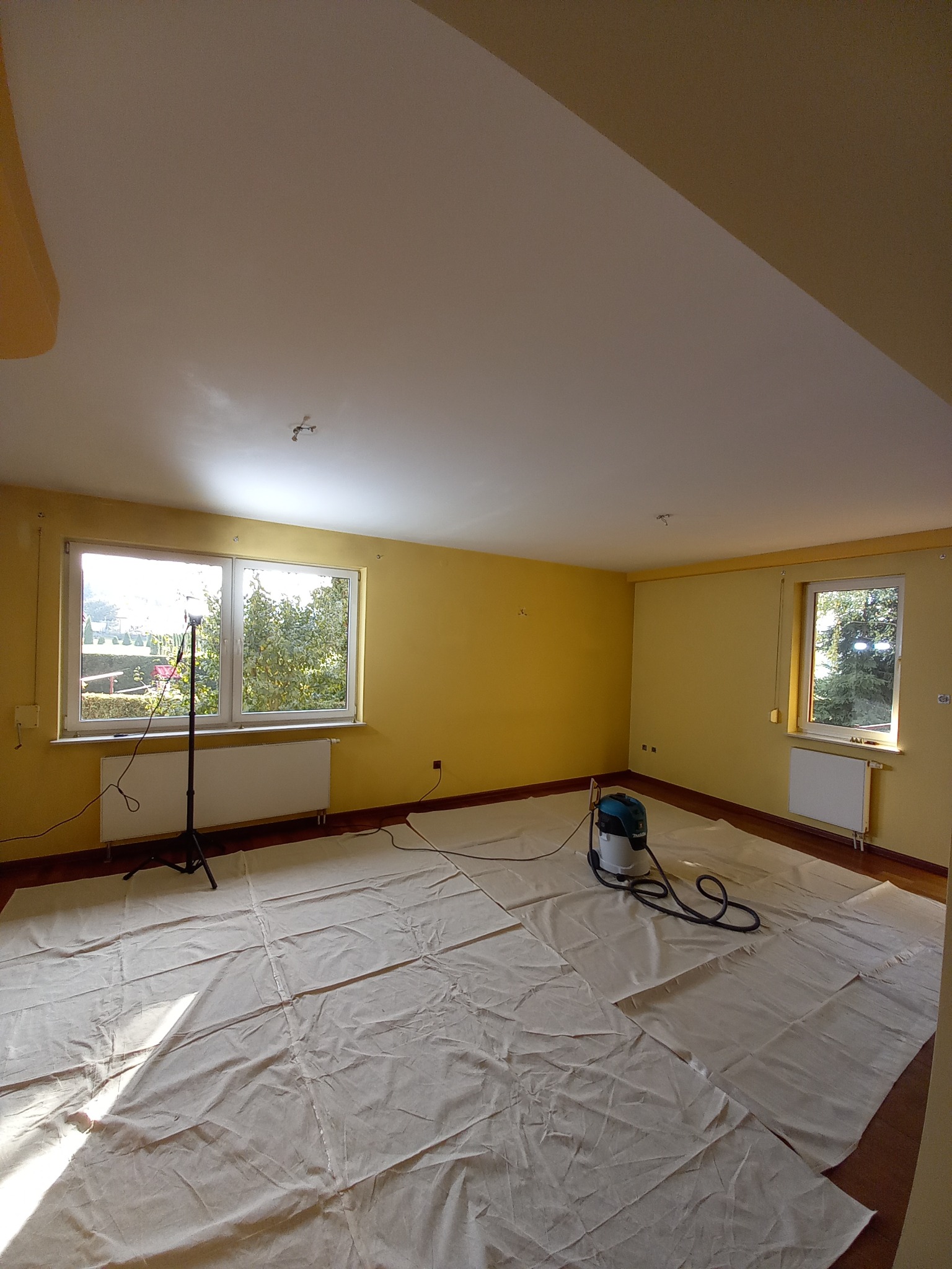 Pokój przygotowany do prac wykończeniowych: żółte ściany, podłoga zabezpieczona płachtami, widoczny odkurzacz przemysłowy Makita i lampa oświetleniowa na statywie.