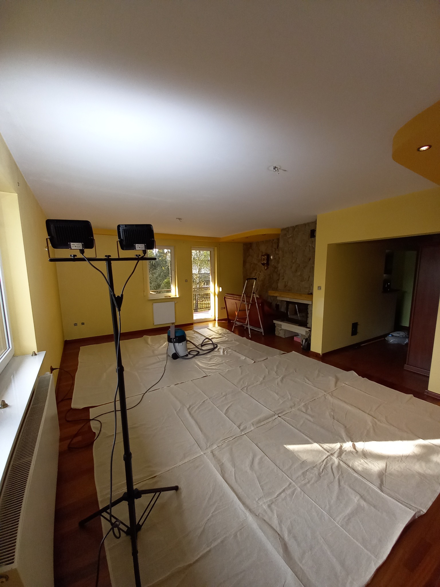 Pokój przygotowany do prac wykończeniowych: podłoga zabezpieczona folią ochronną, ustawione lampy oświetleniowe na statywie, widoczny odkurzacz przemysłowy i drabina przy kamiennym kominku.