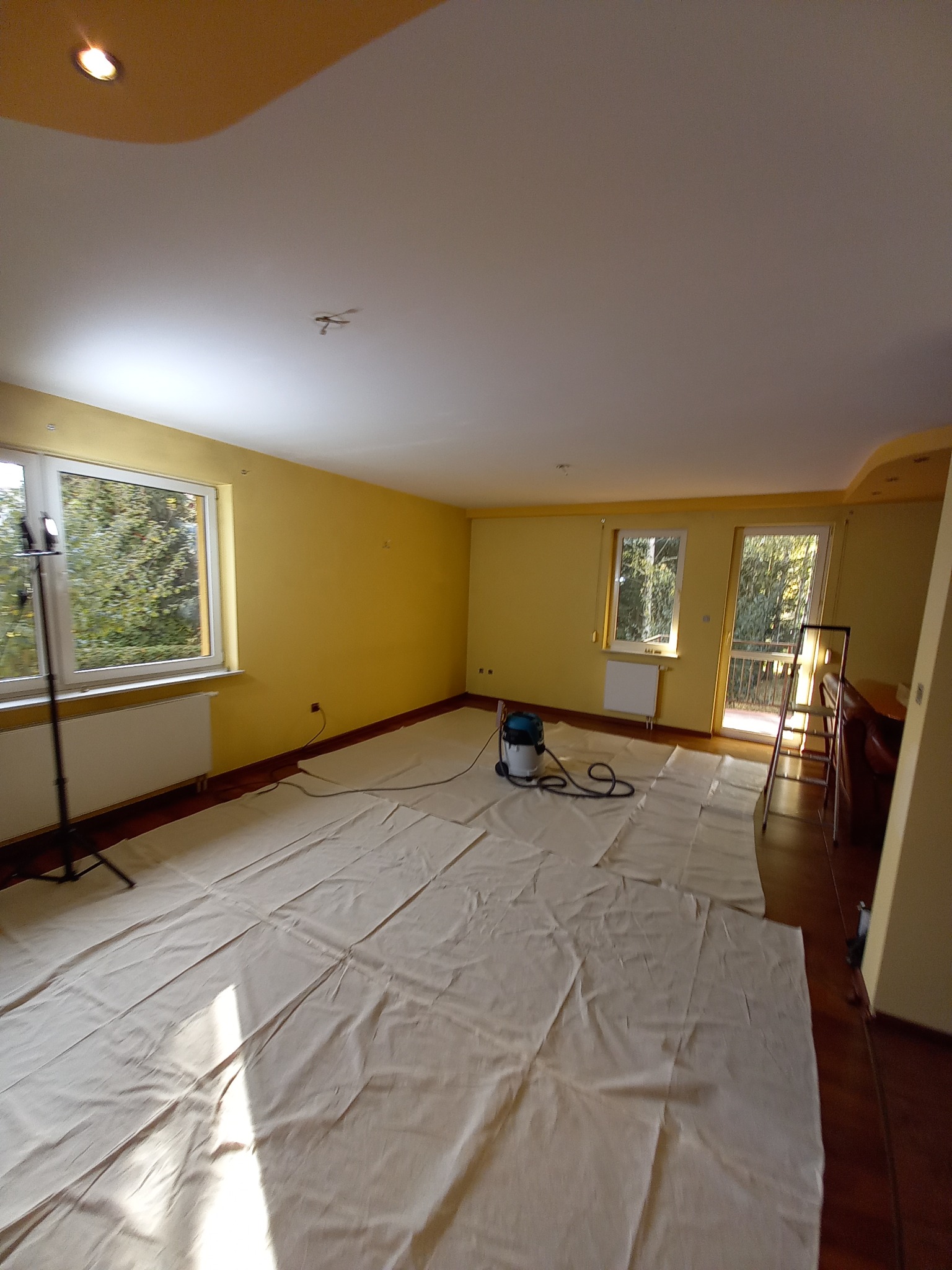 Pokój przygotowany do prac wykończeniowych, z żółtymi ścianami, zabezpieczoną podłogą, widocznym odkurzaczem przemysłowym i drabinką, oświetlony światłem dziennym wpadającym przez okna.