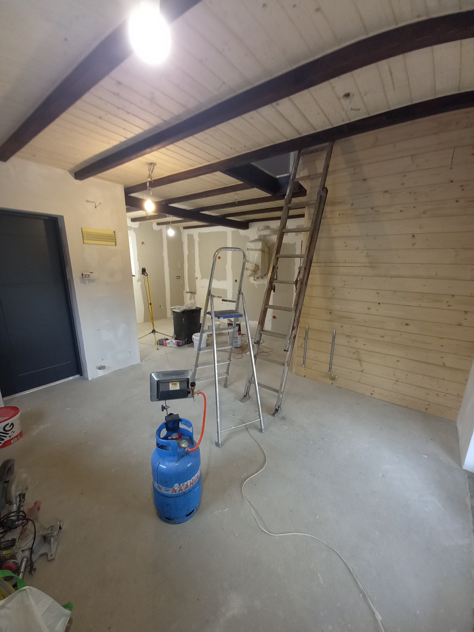Wnętrze w trakcie remontu z drewnianą boazerią, aluminiową drabiną, butlą gazową i nagrzewnicą, widoczne elementy wykończenia sufitu i ściany z płyt kartonowo-gipsowych w tle.