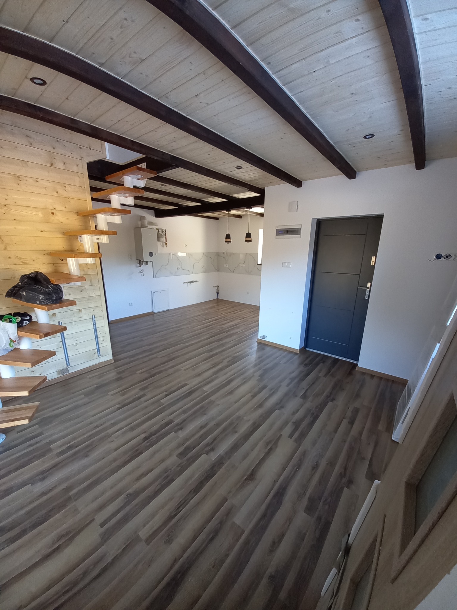 Wnętrze domu po remoncie: drewniana podłoga, białe ściany, drewniany sufit z ciemnymi belkami, schody z drewnianymi stopniami, widoczny fragment kuchni z białymi płytkami imitującymi marmur.