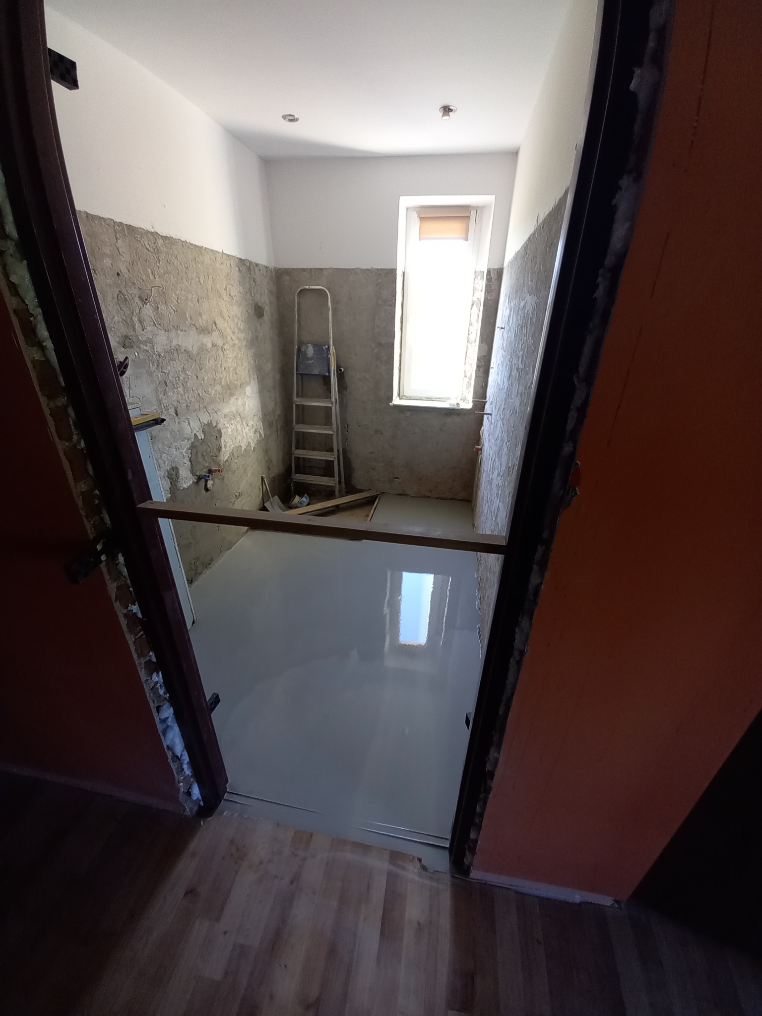 Wnętrze w trakcie prac przygotowawczych do remontu, widoczne ściany z fragmentami starego tynku, nowa wylewka podłogowa, drabina oparta o ścianę przy oknie i surowa ościeżnica drzwiowa.