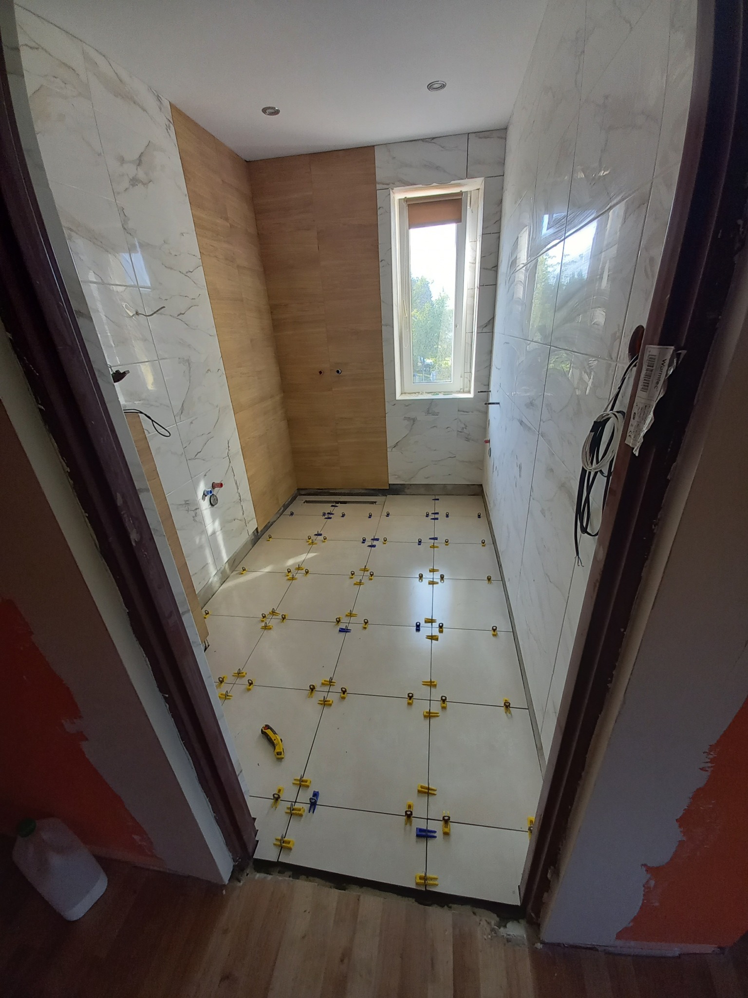 Układanie jasnych płytek podłogowych w łazience, z użyciem żółtych i niebieskich klipsów poziomujących, widoczne płytki na ścianach imitujące marmur oraz drewno, niedokończone wykończenie przy...