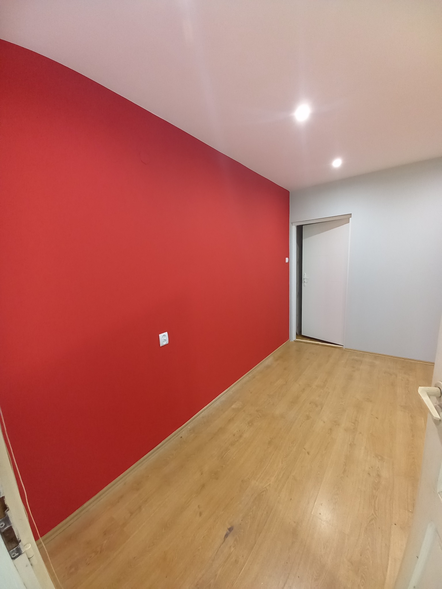 Wnętrze pokoju po remoncie: jedna ściana pomalowana na intensywny czerwony kolor, pozostałe ściany szare, jasna drewniana podłoga, białe drzwi przesuwne w głębi, widoczny kontakt elektryczny.