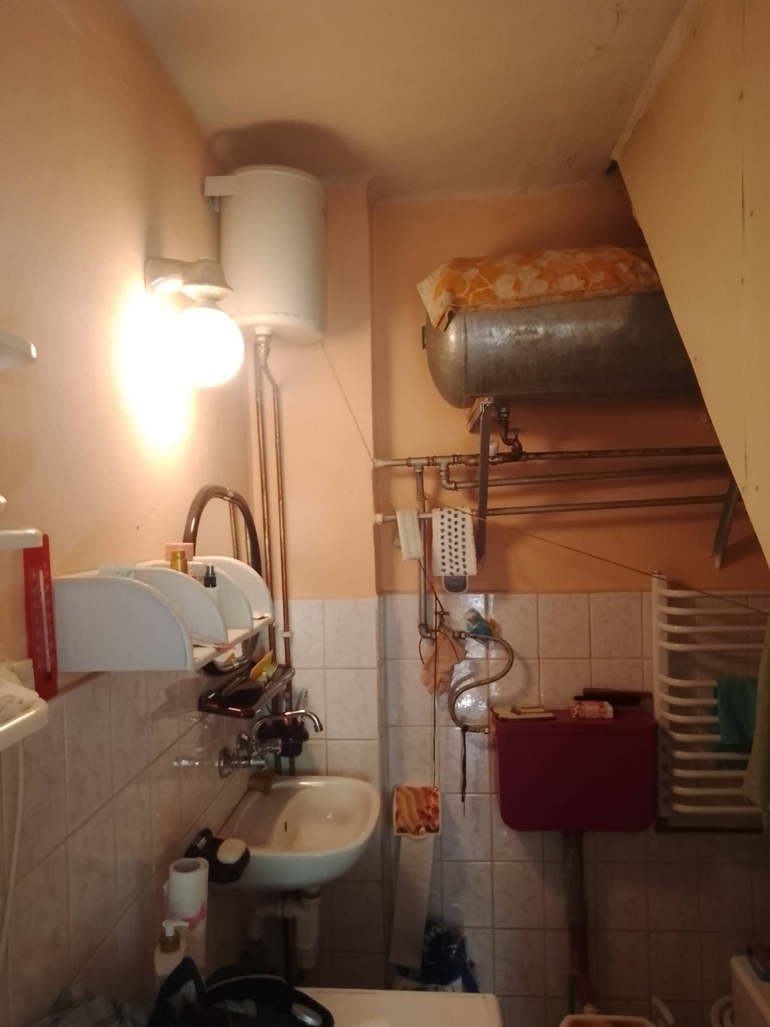 Mała, zaniedbana łazienka z bojlerem elektrycznym i gazowym, ceramicznymi płytkami na ścianach i podłodze, oraz z niezbyt estetycznym wyposażeniem.