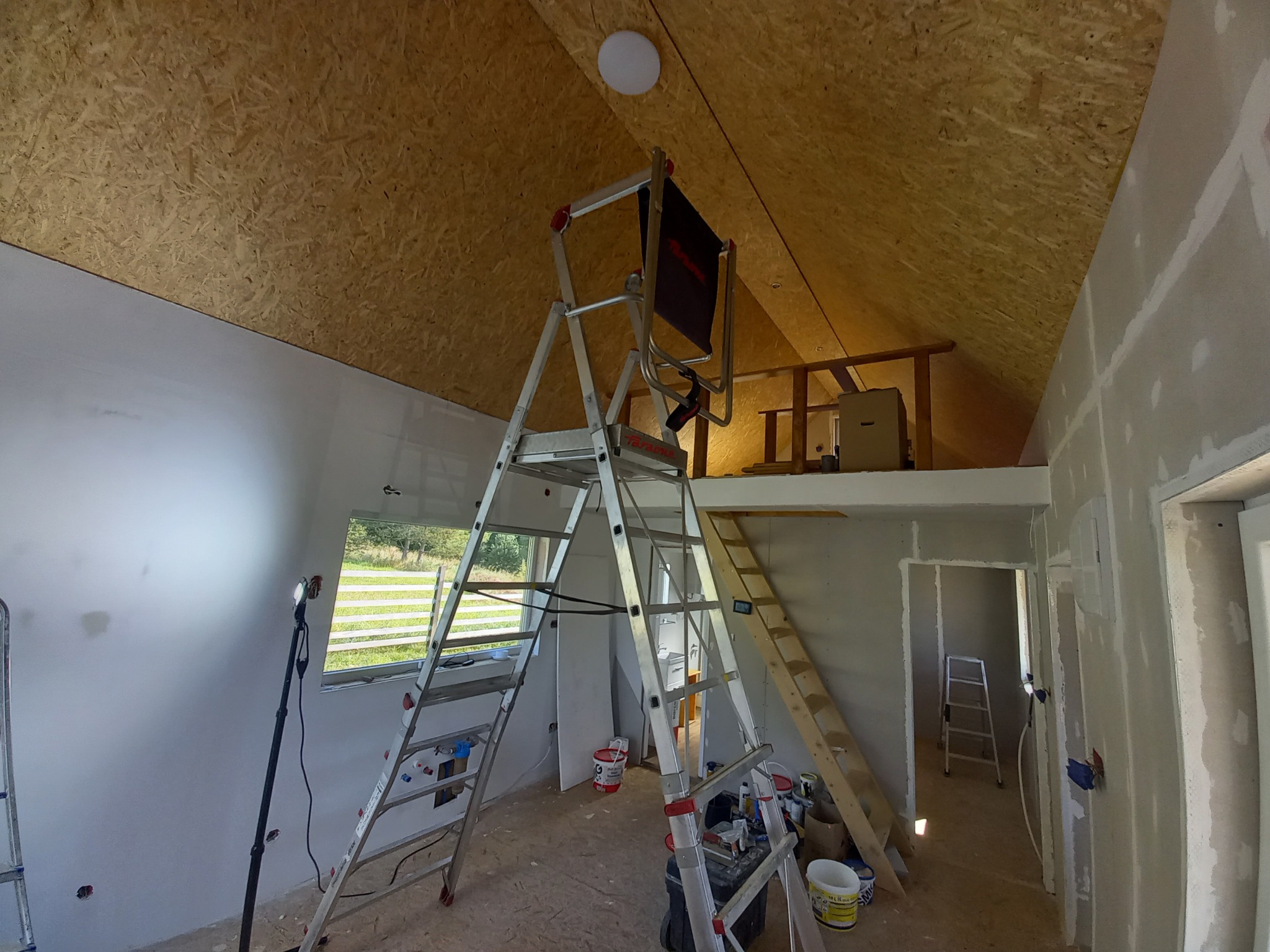 Poddasze w trakcie adaptacji: widoczne ocieplenie drewnem, metalowa drabina z oświetleniem, szpachlowane ściany, okno oraz drewniane schody prowadzące na antresolę.