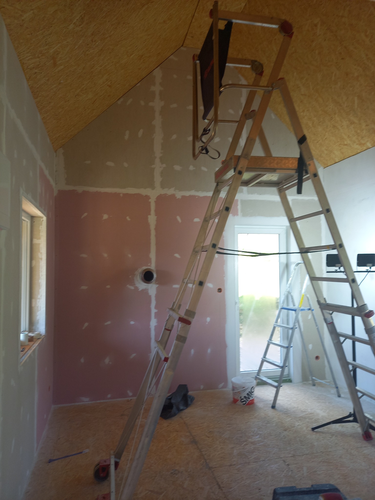 Poddasze w trakcie prac wykończeniowych: widoczne płyty kartonowo-gipsowe na ścianach i suficie, częściowo pokryte gładzią, aluminiowa drabina z podestem na środku pomieszczenia, przygotowanie...