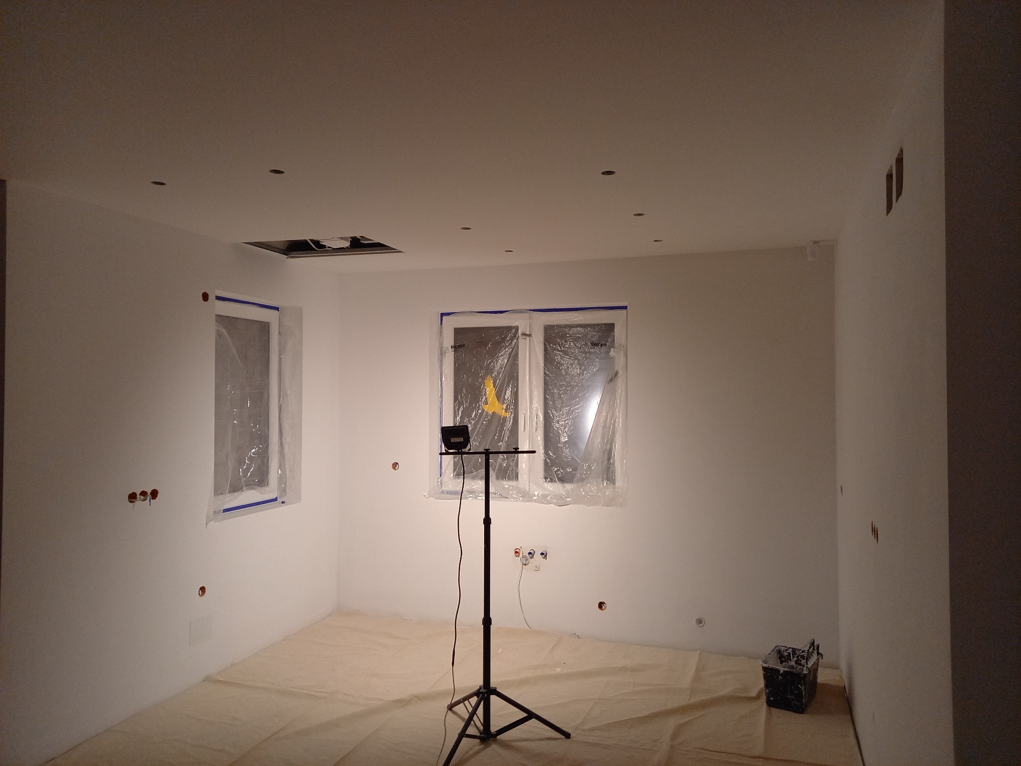Przygotowane do malowania pomieszczenie z zakrytymi folią oknami, zabezpieczoną podłogą, widocznymi puszkami elektrycznymi i lampą na statywie.