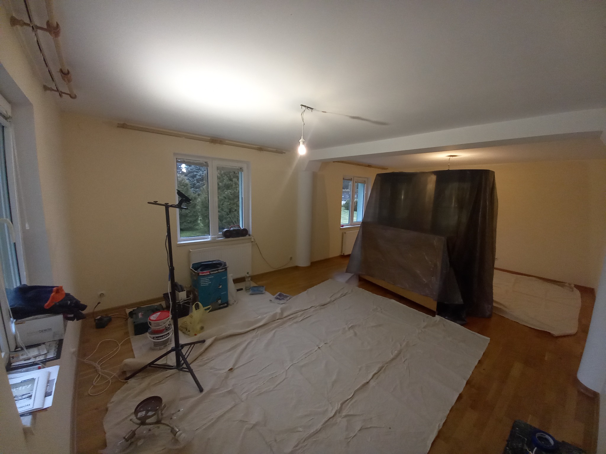 Pokój przygotowany do prac wykończeniowych: podłoga zabezpieczona płachtą malarską, meble przykryte folią, widoczny statyw z oświetleniem, puszki z farbą i narzędzia malarskie.