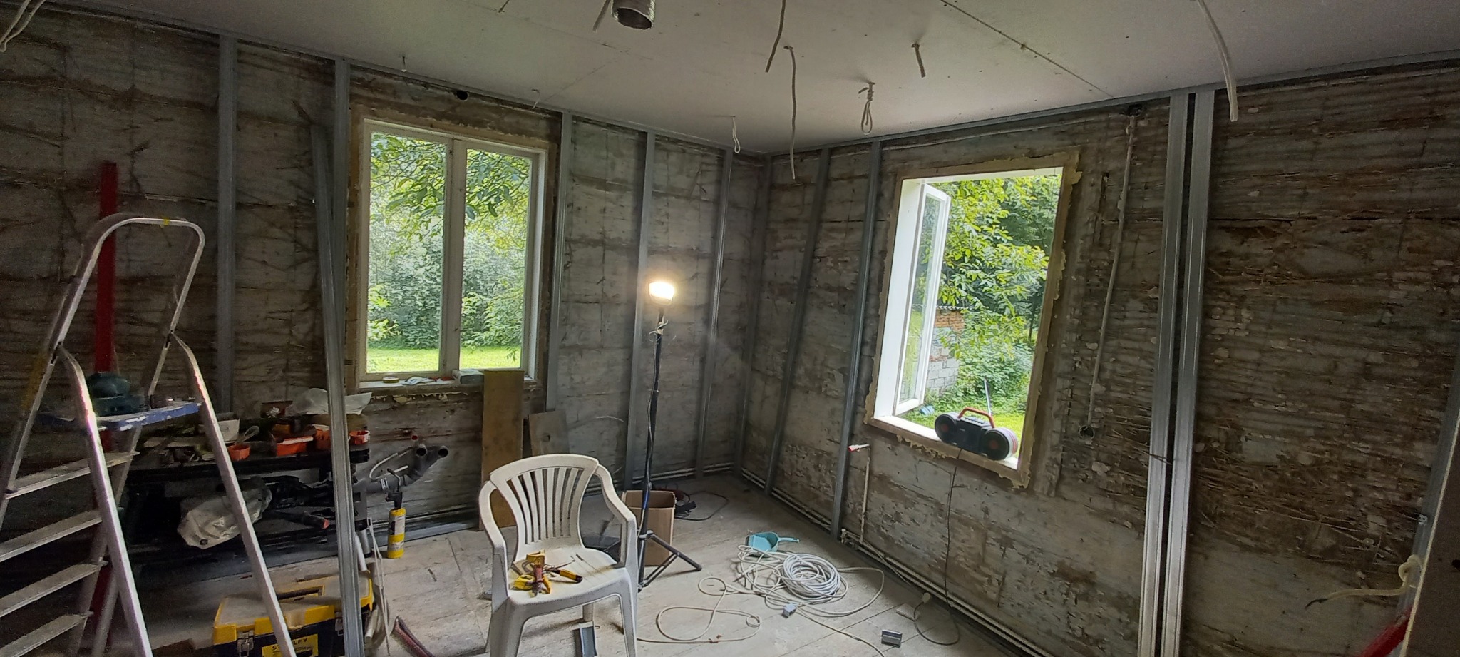 Pokój w trakcie prac przygotowawczych do wykończenia, widoczne metalowe profile na ścianach, odsłonięte fragmenty starej ściany, okna z widokiem na zieleń, drabina, narzędzia i lampa oświetlająca...