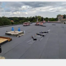 Płaski dach pokryty papą termozgrzewalną z widocznymi kominami, butlami z gazem i narzędziami dekarskimi, częściowo zakończone prace.