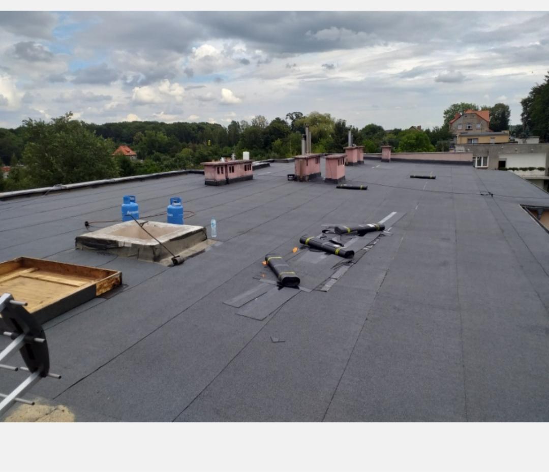 Płaski dach pokryty papą termozgrzewalną z widocznymi kominami, butlami z gazem i narzędziami dekarskimi, częściowo zakończone prace.