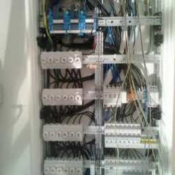 Rozdzielnica elektryczna z bezpiecznikami i przewodami w białej obudowie. Widoczne liczne kable i elementy instalacji.